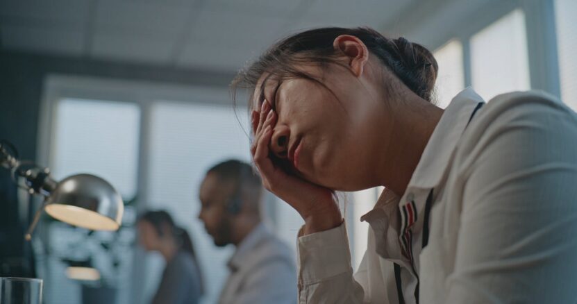 A woman in an office environment looking fatigued, resting her head on her hand while working