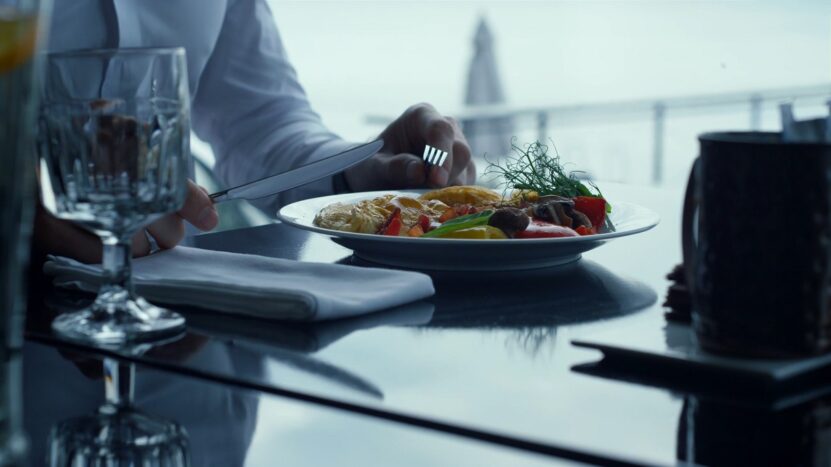 A person in formal attire dining at a restaurant, cutting into a healthy plate of food with vegetables and fish, with a scenic outdoor view in the background